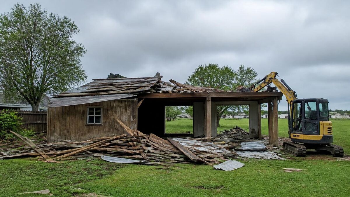 A storm-damaged shed and carport being torn down in a flat backyard with a small excavator