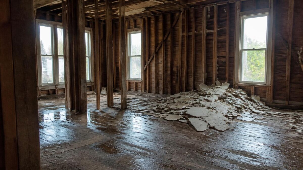 The interior of an older raised house gutted to the framing, with plaster and drywall debris on the wood floor