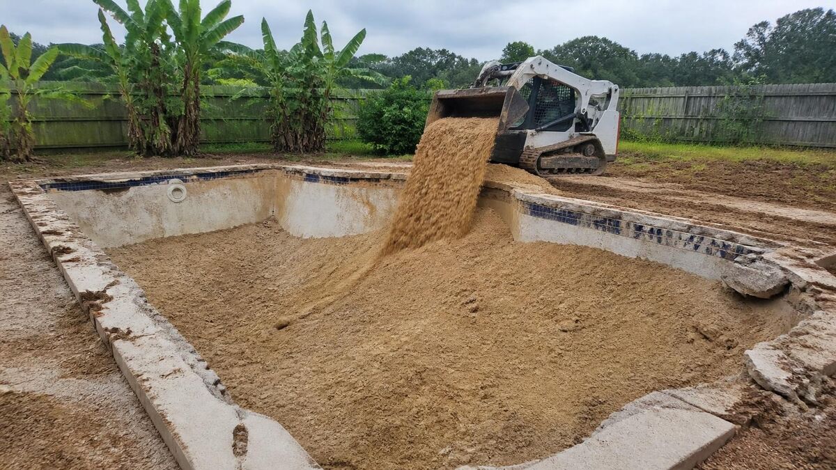 A small loader filling an old in-ground pool with sand and dirt in a flat backyard